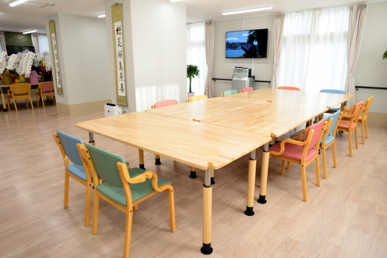 明るい共用スペース。大きな木製テーブルとカラフルな椅子、壁掛けテレビ、生花が配置されている。